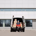 Man in a colorful jacket sitting by a van with boxes in an urban setting.